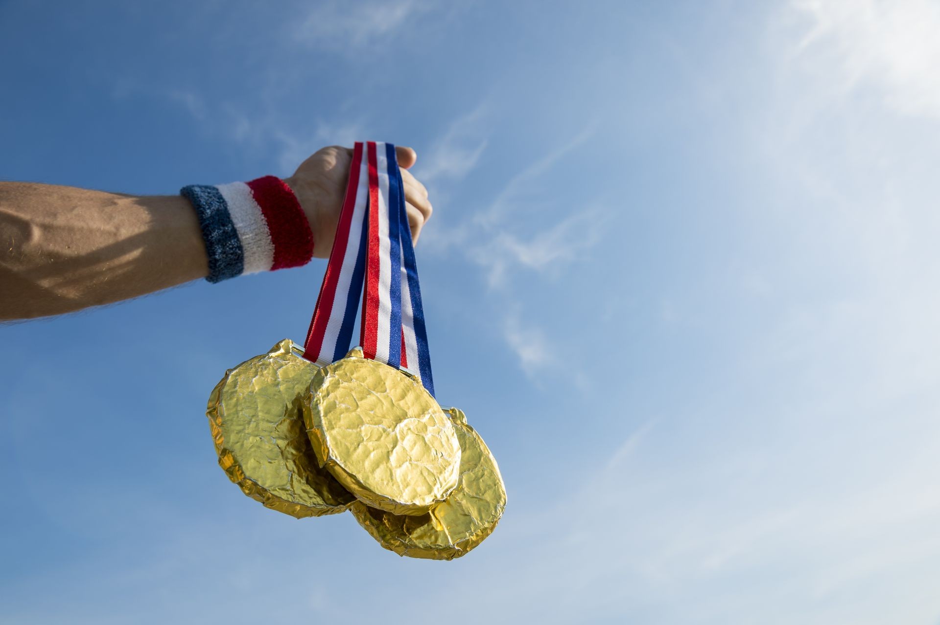 Hand of American athlete holding gold medals hanging from USA colors red, white, and blue ribbon against a bright blue sky background. Backlit with lens flare.